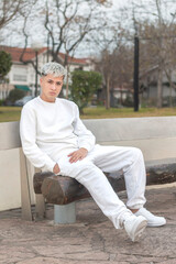 Joven rubio sentado tranquilamente en una plaza urbana, vestido completamente de blanco. La imagen captura un momento de serenidad y relajación al aire libre.