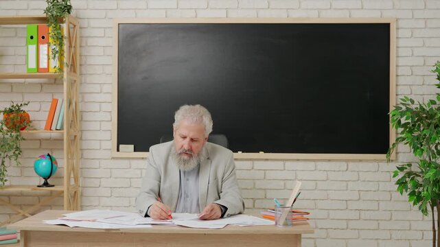 Aged man college professor sit at desk in classroom in front of chalkboard grading test papers with red marker. Education concept.