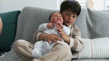 Young boy holding newborn baby with thoughtful expression on a sofa. baby looks up contentedly, creating a heartwarming scene of sibling bonding and family love in a cozy home setting