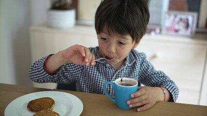 Young boy in checkered shirt enjoying a hot drink and pancakes, smiling and playfully licking the spoon, seated at a wooden table in a cozy home environment