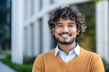 Confident young professional with curly hair and warm smile standing outdoors near modern office building. Image conveys success, positivity, and approachable demeanor in business context