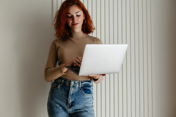 Redheaded Woman Holding Laptop, Curly Hair Framing Face, Against a Light-Colored Wall