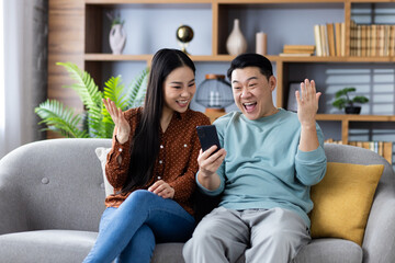 Asian family enjoying exciting news on a phone while sitting on a sofa at home. Mature couple...