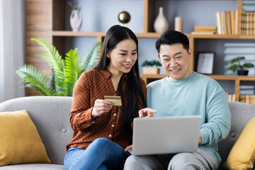 Happy Asian family mature couple sitting on couch using laptop and credit card for online shopping. Both smiling and engaged in activity, depicting love, technology, and home comfort.