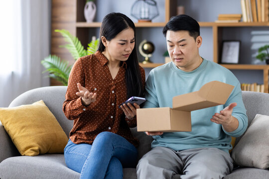 Mature Asian couple sitting on living room couch looking confused with online order. Man holding open package, woman holding smartphone. Scene depicts frustration, modern technology - Powered by Adobe