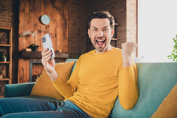 Photo portrait of young guy hold device winning raise fist sit couch dressed yellow garment spend...
