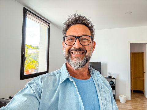 Handsome adult man with a beard and glasses taking a selfie in his new apartment. At 50 years old, he captures the joy of settling into a modern home, reflecting a new chapter and achievement.