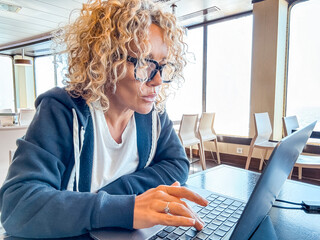 Casual adult woman with curly hair and glasses working remotely on her laptop at a desk aboard a...