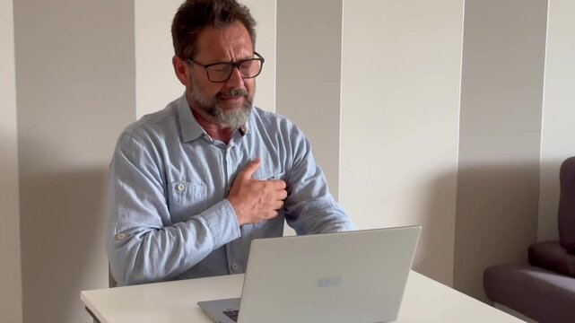 Chest pain. Bearded middle-aged man with glasses having heart attack. Computer user with stress experiencing chest pain. Heart attack while working on laptop at home office desk