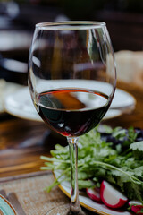 Glass of red wine, it shows a close-up of a glass of red wine sitting on a table, against a backdrop of greens and salad