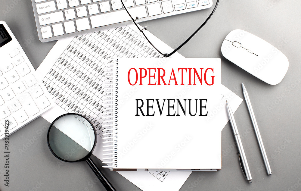 Poster notebook with the words operating revenue on a desk with a keyboard, calculator, pen, and charts