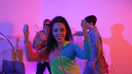Attractive happy dancer looking at camera while smart woman moving at rhyme with neon light. Professional hispanic performer break dancing while wearing colorful cloth with diverse friend. Regalement.