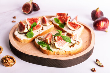 Tasty homemade sandwiches with sweet figs, cream cheese, walnuts. honey and mint leaves on the wooden serving plate on light marble background close up.