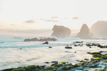 Serene coastal landscape at sunrise with moss-covered rocks and misty waves crashing against unique rock formations. Perfect for tranquil, nature-inspired projects. - Powered by Adobe