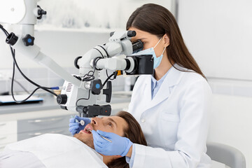 Female professional dentist doctor making treatment in modern cabinet, using microscope