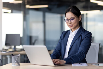 Asian businesswoman working at laptop in modern office, focused and confident, demonstrating professional skills and dedication.