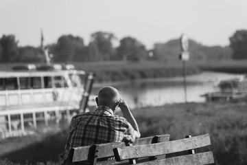 an elderly man sits thoughtfully on a bench by the river, illuminated by sunlight, black and white photo