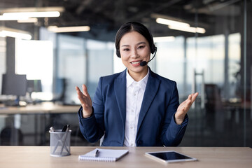 Asian businesswoman wearing headset smiling, talking on video call in modern office, connected with phone and tablet.