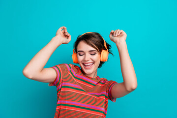 Portrait of funky positive girl with short hairstyle wear stylish t-shirt in headphones raising hands up isolated on teal color background