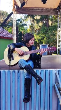 A small, beautiful girl in jeans and a leather jacket plays a six-string acoustic guitar, alone on the street