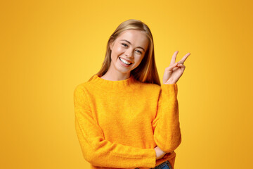 Smiling redhead teenager showing peace gesture and posing to camera, orange studio background with free space
