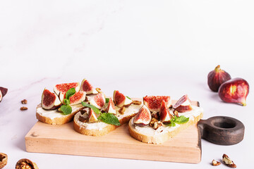 Tasty homemade sandwiches with sweet figs, cream cheese, walnuts. honey and mint leaves on the wooden serving plate on light marble background close up.