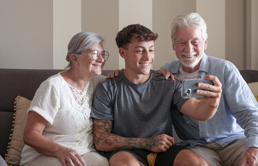 Happy multigenerational family of white-haired grandparents and young grandson taking selfie with cell phone sitting together at home. Smiling three generations of caucasian family