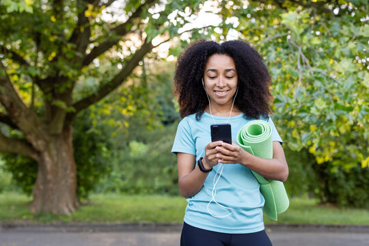 Smiling woman in park with yoga mat and phone, wearing earphones, enjoying fresh air and fitness motivation. Relaxed and ready for a workout.