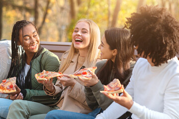 Cheerful multiracial teens joking and laughing while eating pizza, sitting on bench in park