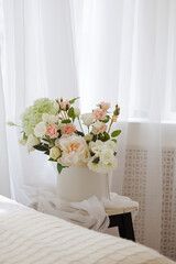 A white vase with pink flowers sits on a wooden stool. The flowers are arranged in a way that creates a sense of balance and harmony. The vase and stool are placed on a bed