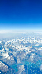 Obraz premium Out of this World Views Flying Over Greenland. Snow-capped mountains, glaciers and pristine terrain. Clear blue skies above the horizon.