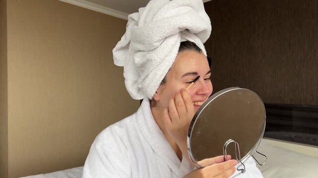 Horizontal 4K video. Young adult white woman in robe, towel on head, looks into small round mirror, combs eyebrows in cozy hotel bed room. Cosmetic procedure in spa salon, rest, relaxation, march 8, 