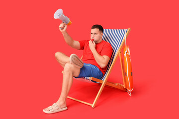 Young lifeguard with lifebuoy and megaphone sitting on beach lounge and whistling against red...