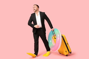 Happy office worker in flippers with inflatable ring and suitcase on pink background