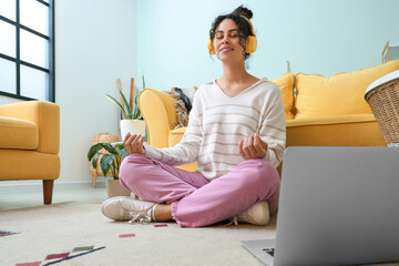 Beautiful young happy African-American woman in headphones with laptop meditating at home