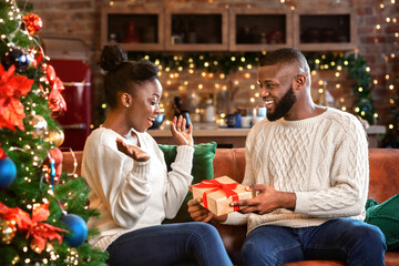 Christmas morning. African american couple celebrating Xmas at home together, handsome man giving present to his delighted wife