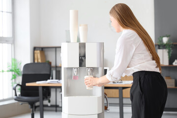 Businesswoman pouring water from cooler in office, back view