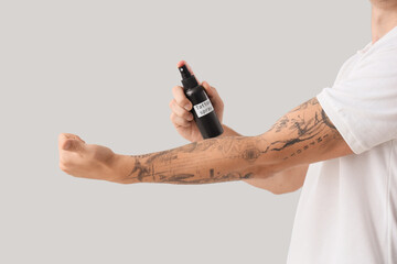 Young man spraying his tattooed arm on light background, closeup
