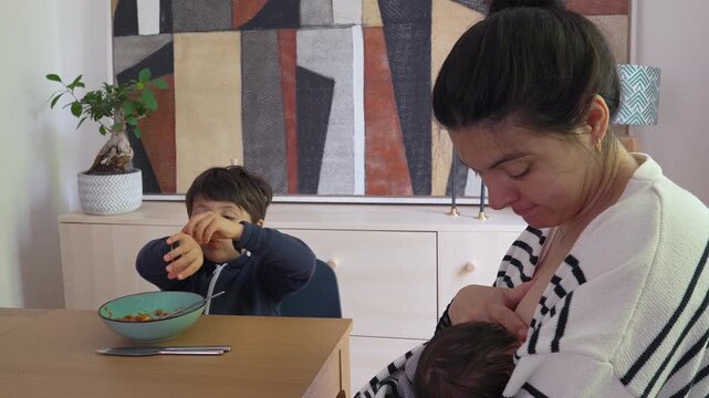 Mother breastfeeding newborn while older son sits at the dining table, seemingly distracted during mealtime, representing the everyday juggle of parenting two young children