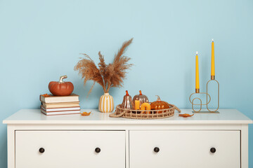 Decorative pumpkins with burning candles, pampas grass and books on commode near blue wall
