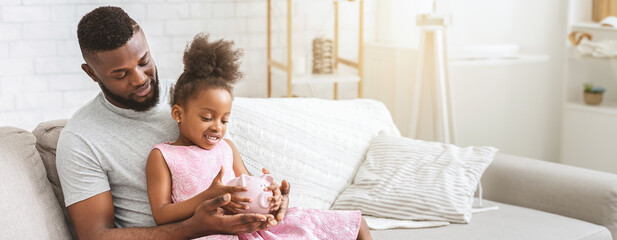 Black father and daughter share a joyful moment on a cozy couch, as the little girl excitedly holds...