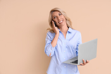 Mature businesswoman with laptop talking by mobile phone on beige background