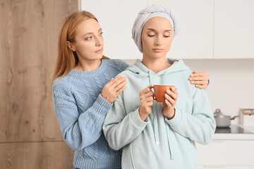 Sad young woman after chemotherapy with coffee cup and her mother in kitchen