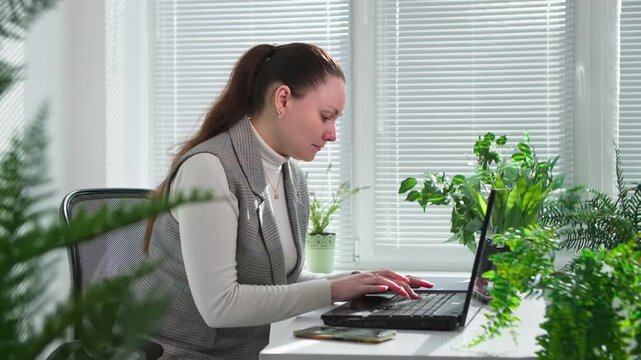 cheerful atmosphere in office, young woman works on laptop and covers herself with folder from flying boulders in office with green plants