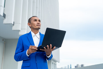 A confident businessman in a blue suit holding a laptop outdoors, gazing upwards, symbolizing ambition, forward-thinking, and strategic vision in a modern urban environment.