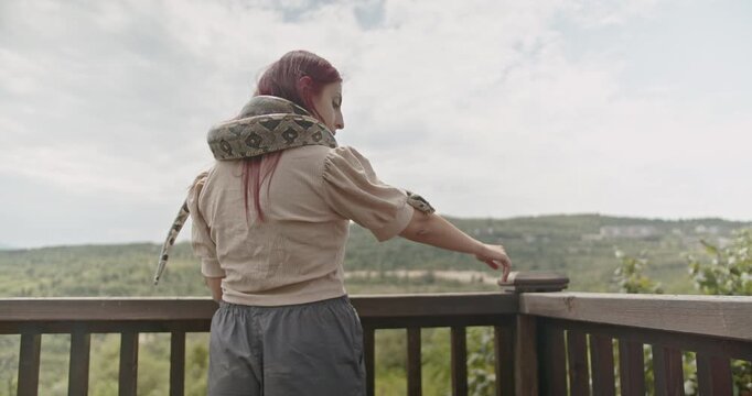 A young woman stands on a balcony with her pet snake wrapped around her shoulders.