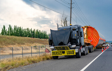 Naklejka premium Broken black big rig semi truck with open hood and oversized load on semi trailer standing out of service on road shoulder waiting for road assistant help and repair