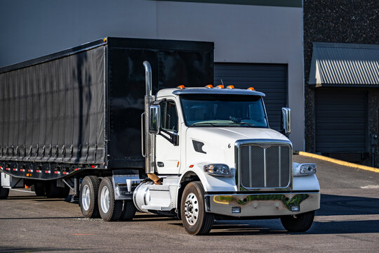 Powerful day cab white big rig semi truck with black Conestoga flat bed semi trailer standing in warehouse dock loading cargo for next freight
