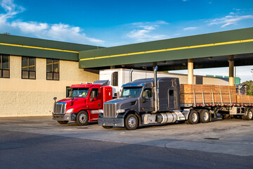 Two different long haul red and gray big rigs semi trucks with loaded refrigerator and flat bed semi trailers standing on truck stop parking lot take a break