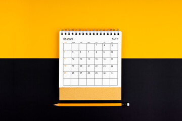 Top view of May 2025 desk calendar and pencil on yellow and black background.